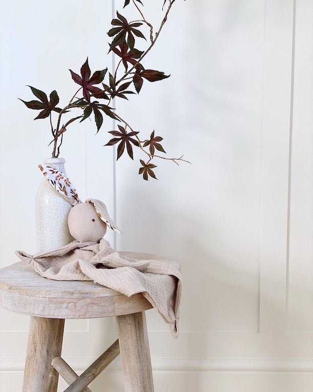 Decorative setup with a vase, plant, and small figurine on a wooden stool against a white paneled wall.